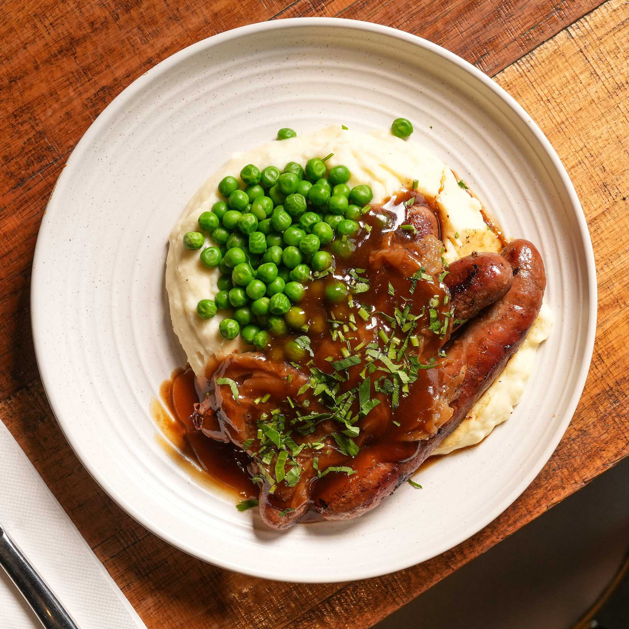 Smokehouse Bangers & Mash | Beef Sausages Served Over Creamy Mash With Rich Onion Gravy And Peas | $30.80